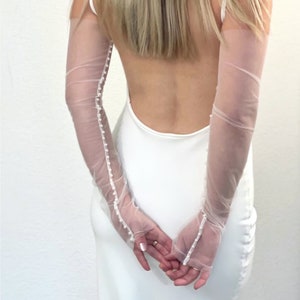Könnte beinhalten: Weiße, durchsichtige Handschuhe mit einer Reihe von Perlen, die auf der Rückseite jedes Handschuhs angenäht sind. Die Handschuhe werden von einer Person mit langen blonden Haaren getragen. "Stitched Pearl Seam" steht in einem weißen Rechteck oben im Bild.