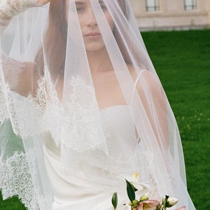 May include: A bride wearing a white wedding dress with a lace trim veil and holding a bouquet of white and pink flowers.