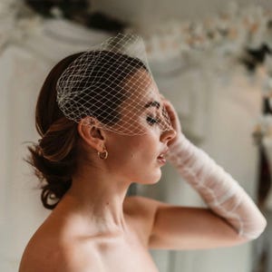 May include: A bridal veil with a net design, covering the forehead and eyes. The veil is white and paired with a matching long glove. The model is wearing gold hoop earrings. The overall theme is bridal fashion.