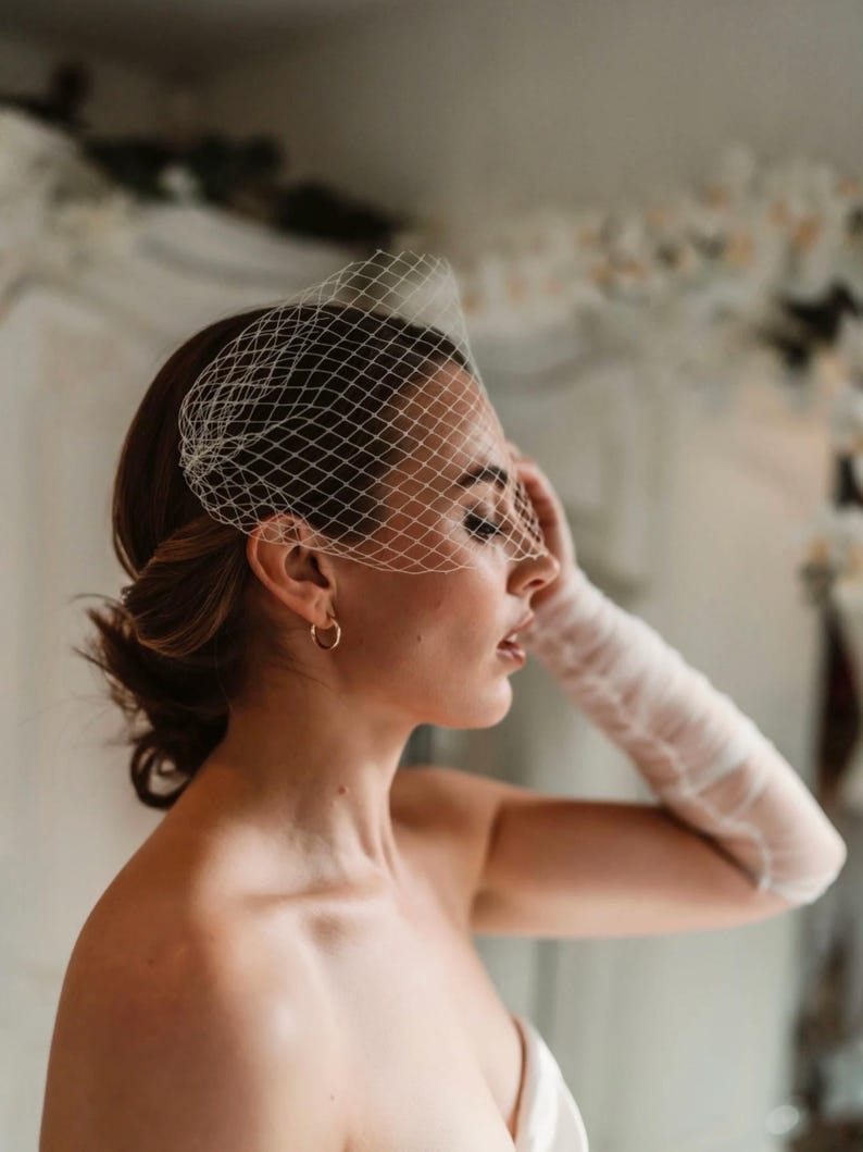 May include: A bridal veil with a net design, covering the forehead and eyes. The veil is white and paired with a matching long glove. The model is wearing gold hoop earrings. The overall theme is bridal fashion.