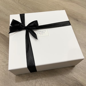 K&ouml;nnte beinhalten: Eine wei&szlig;e Geschenkbox, gebunden mit einem schwarzen Satinband und einer Schleife. Auf der Box befindet sich ein Etikett mit der Aufschrift "HARRIETT FALVEY - FASHION DESIGNER". Auf hellem Holzgrund.