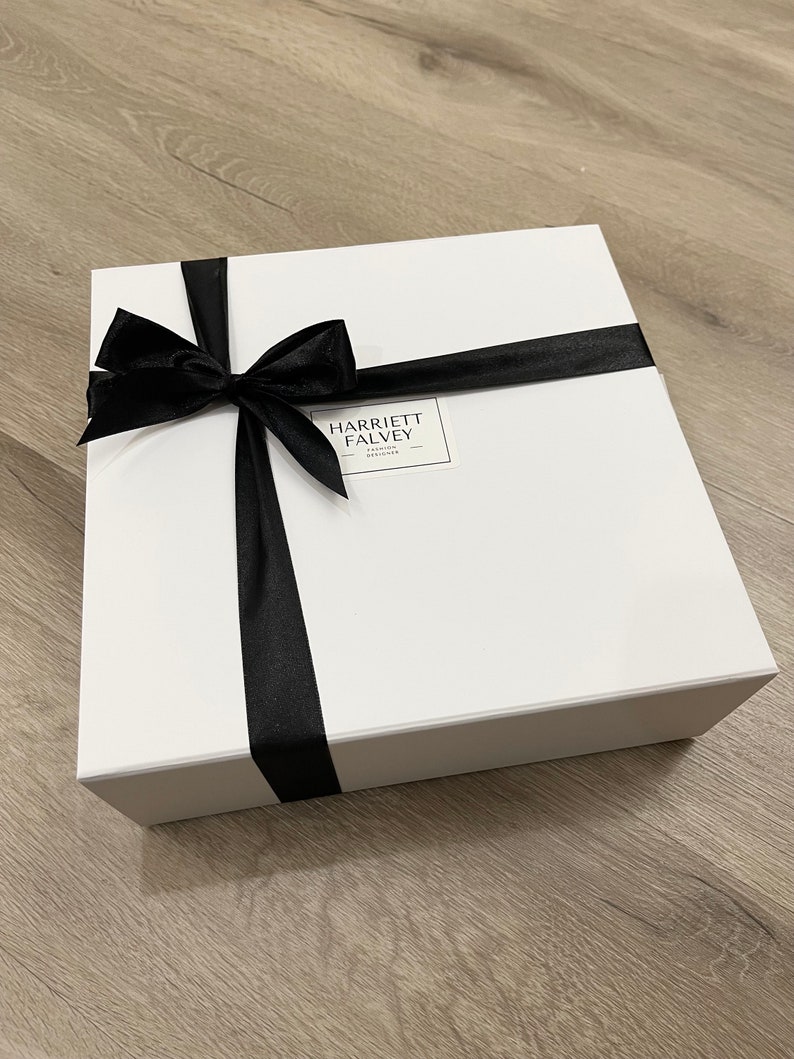 K&ouml;nnte beinhalten: Eine wei&szlig;e Geschenkbox, gebunden mit einem schwarzen Satinband und einer Schleife. Auf der Box befindet sich ein Etikett mit der Aufschrift "HARRIETT FALVEY - FASHION DESIGNER". Auf hellem Holzgrund.