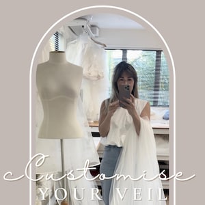 Pode incluir: Uma mulher com uma blusa branca e calças jeans azuis está em pé em uma sala com um manequim branco e um véu branco drapeado sobre ele. O texto "Customise Your Veil" está sobreposto à imagem.