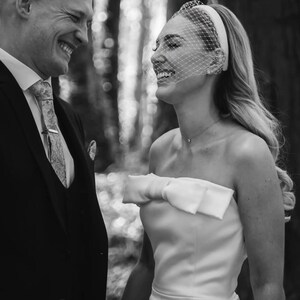 May include: Black and white photo of a smiling couple. The woman wears a strapless white dress with a bow. She has a veil and headband. The man wears a suit and tie. Text reads "Bride Chloe by @infiniteimages_."
