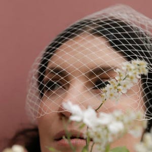 May include: A close-up of a person wearing a white net veil. White flowers are in the foreground, with a pink background. Text at the top reads "Bride Tani by @melodyjoyco as seen in Tatler magazine."
