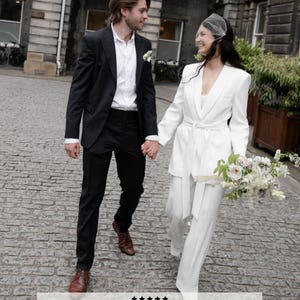 May include: A couple walking on a cobblestone street, the woman in a white pantsuit and veil, holding a bouquet. The man wears a black suit. Text: "Loved my veil so much, thank you Harriett for your incredible work!" Bride Tani by @melodyjoyco.