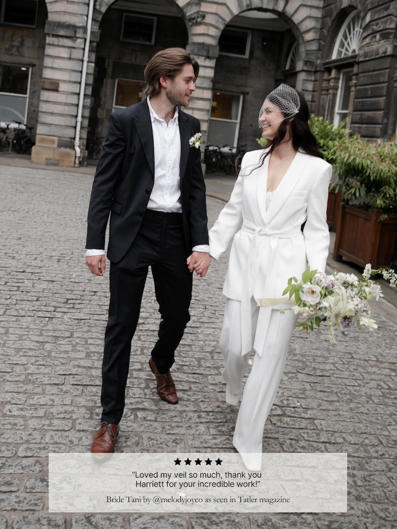 May include: A couple walking on a cobblestone street, the woman in a white pantsuit and veil, holding a bouquet. The man wears a black suit. Text: "Loved my veil so much, thank you Harriett for your incredible work!" Bride Tani by @melodyjoyco.