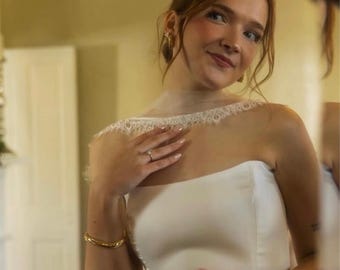 Op de afbeelding: Een strapless witte trouwjurk met een glad, effen lijfje en een delicate, doorschijnende witte sluier met een verfijnde kanten rand. De sluier is over de schouders gedrapeerd, wat het gedetailleerde bloemmotief van de kanten rand benadrukt.