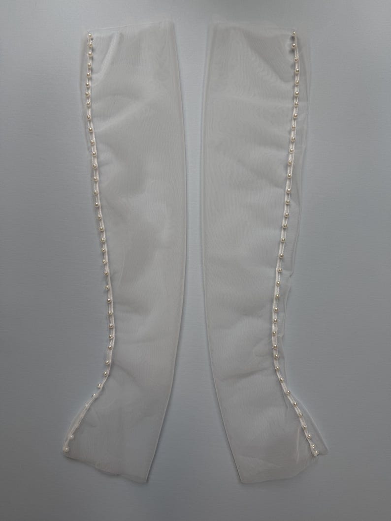 May include: A pair of long, sheer white gloves made of a translucent mesh fabric. Each glove features a vertical row of small, round, white faux pearl buttons running along the outer edge from the wrist to the top opening of the garment.