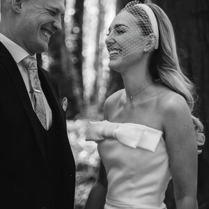 May include: Black and white photograph of a smiling couple. The woman wears a strapless white wedding dress with a large bow. She has a veil and headband. The man wears a black suit and patterned tie. Text at the bottom reads "Bride Chloe by @infiniteimages_."