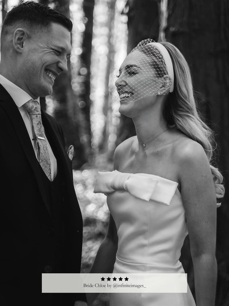 May include: Black and white photograph of a smiling couple. The woman wears a strapless white wedding dress with a large bow. She has a veil and headband. The man wears a black suit and patterned tie. Text at the bottom reads "Bride Chloe by @infiniteimages_."