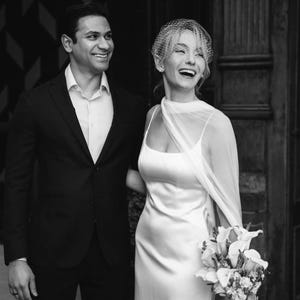 May include: Black and white photo of a smiling couple on their wedding day. The bride wears a white satin gown and veil, holding a bouquet. The groom is in a black suit. Text at the bottom reads "Bride Kelsey by @lennypellico".