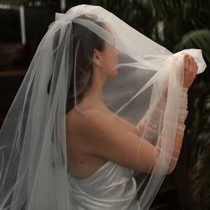 Könnte beinhalten: Eine Braut in einem weißen Satin-Hochzeitskleid, die einen durchsichtigen Schleier hält. Der Schleier fällt über den Kopf und die Schultern der Braut und erzeugt einen weichen, ätherischen Effekt. Die Ärmel sind aus dem gleichen transparenten Material.