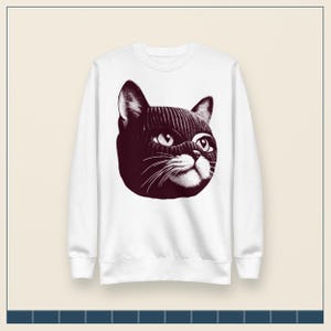 May include: A white long-sleeve crewneck sweatshirt featuring a large, dark maroon graphic print on the chest. The graphic depicts a cat wearing a ribbed knit balaclava, showing only its eyes, nose, and whiskers in a detailed, stippled art style.
