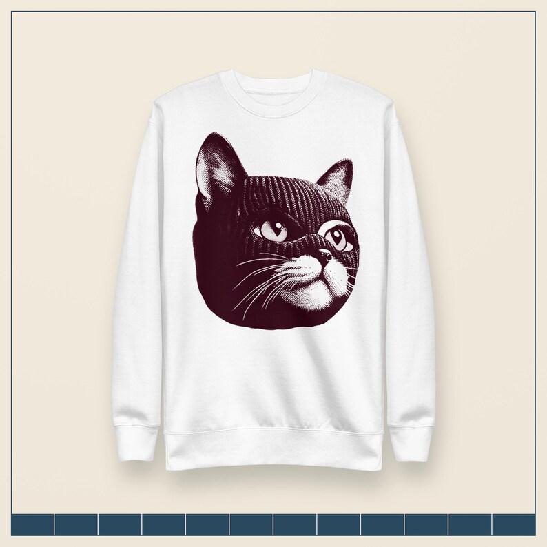 May include: A white long-sleeve crewneck sweatshirt featuring a large, dark maroon graphic print on the chest. The graphic depicts a cat wearing a ribbed knit balaclava, showing only its eyes, nose, and whiskers in a detailed, stippled art style.