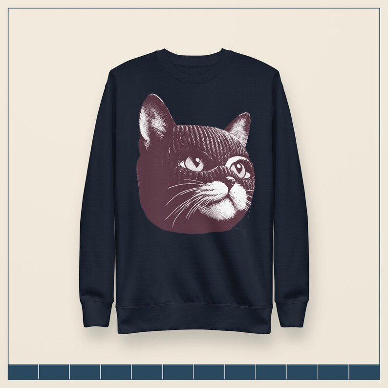 May include: A dark navy blue long-sleeve sweatshirt featuring a large, centered graphic illustration of a cat wearing a dark-colored knit balaclava. The cat's face is rendered in a detailed, high-contrast style with white and dark maroon tones.