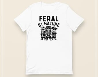 Puede incluir: Una camiseta blanca de manga corta con un estampado gráfico negro de tres gatos con gafas de sol, gorras de béisbol y cadenas de oro. Sobre los gatos, el texto dice FERAL BY NATURE en una fuente negra, negrita y sin remates.