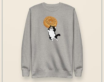 May include: A heather gray crewneck sweatshirt featuring a graphic illustration of a black and white cat standing on its hind legs while holding a large, golden-brown bagel with visible seeds above its head with both front paws.