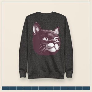 May include: A dark charcoal gray long-sleeve sweatshirt featuring a graphic print of a cat wearing a dark purple knit balaclava. The cat illustration is rendered in a detailed, etched style with white highlights on the eyes, whiskers, and muzzle.