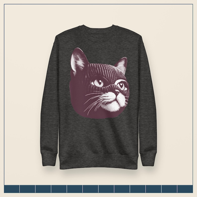 May include: A dark charcoal gray long-sleeve sweatshirt featuring a graphic print of a cat wearing a dark purple knit balaclava. The cat illustration is rendered in a detailed, etched style with white highlights on the eyes, whiskers, and muzzle.