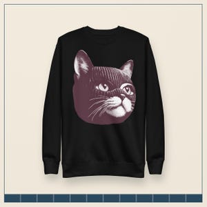 May include: A black long-sleeve sweatshirt featuring a graphic print of a cat wearing a dark-colored knit balaclava. The cat illustration is rendered in a stippled, monochromatic style, highlighting the feline's eyes, whiskers, and nose.