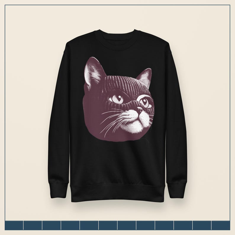May include: A black long-sleeve sweatshirt featuring a graphic print of a cat wearing a dark-colored knit balaclava. The cat illustration is rendered in a stippled, monochromatic style, highlighting the feline's eyes, whiskers, and nose.