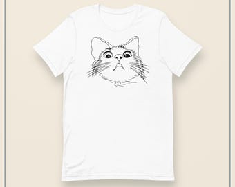 Könnte beinhalten: Ein weißes Kurzarm-T-Shirt mit einer schwarzen Strichzeichnung eines Katzengesichts mit weit aufgerissenen Augen und Schnurrhaaren, die mittig auf der Brust platziert ist. Das Design ist in einem einfachen Skizzenstil auf weißem Stoff gehalten.