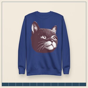 May include: A royal blue long-sleeve sweatshirt featuring a large, centered graphic illustration of a cat wearing a dark-colored knit balaclava. The cat is depicted in a stylized, high-contrast white and dark brown print with visible whiskers.