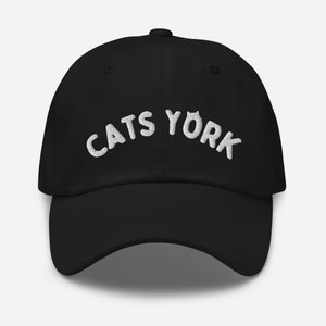Op de afbeelding: Zwarte baseballpet met witte geborduurde tekst die "CATS YORK" in een boogvormige vorm leest.