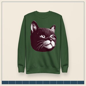 May include: A forest green long-sleeve sweatshirt featuring a large, centered graphic illustration of a cat wearing a dark-colored knit balaclava. The cat's face is rendered in high-contrast white and dark tones, highlighting its eyes and whiskers.