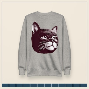 May include: A heather gray crewneck sweatshirt featuring a dark maroon graphic print of a cat wearing a knitted balaclava. The cat illustration shows detailed eyes, whiskers, and a textured knit pattern on the head covering, centered on the chest.