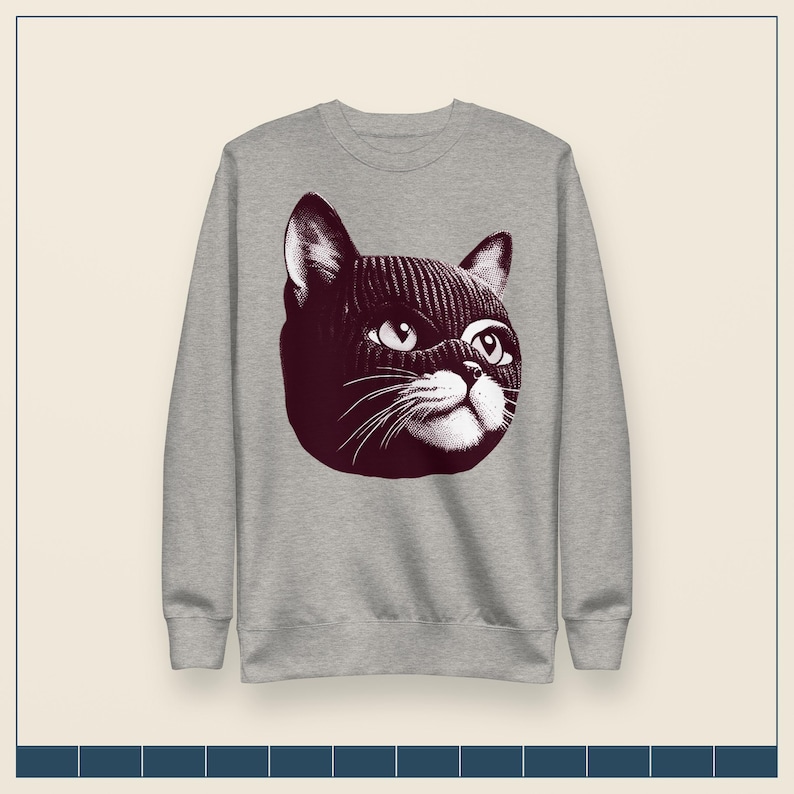 May include: A heather gray crewneck sweatshirt featuring a dark maroon graphic print of a cat wearing a knitted balaclava. The cat illustration shows detailed eyes, whiskers, and a textured knit pattern on the head covering, centered on the chest.