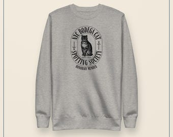 May include: A heather gray crewneck sweatshirt featuring a black graphic print of a cat centered within an oval frame. The text reads NYC BODEGA CAT SPOTTING SOCIETY SINCE 1824 HONORARY MEMBER. The garment has long sleeves and a ribbed neckline.