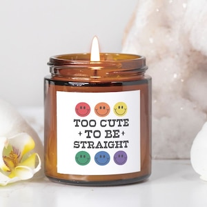 May include: A brown glass candle jar with a white label that reads "Too Cute + To Be + Straight" with rainbow smiley faces. The candle is lit and surrounded by white orchids and a white crystal.