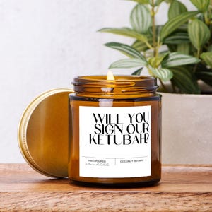 May include: A brown glass candle with a white label that reads "WILL YOU SIGN OUR KETUBAH?"  The candle is hand-poured with coconut soy wax.