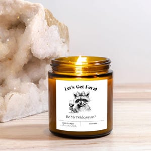 May include: A lit soy wax candle in a brown glass jar. The label features a raccoon illustration holding a wine glass, with the text "Let's Get Feral. Be My Bridesman?" and "Hand Poured". The candle is on a light wood surface.