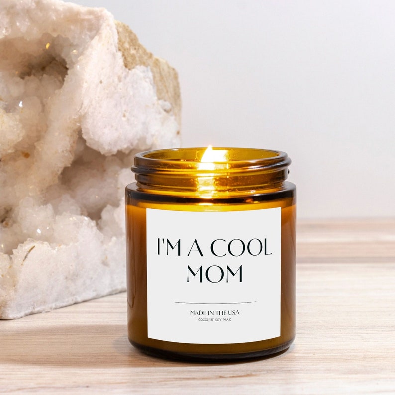 May include: A brown glass candle with a white label that reads "I'm a Cool Mom" and "Made in the USA Coconut Soy Wax". The candle is lit and the flame is visible.