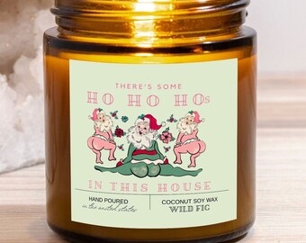 May include: A brown glass candle jar with a white label that reads "There's some Ho Ho Hos in this house". The label features a cartoon illustration of three Santa Clauses with the middle Santa sitting and the two on the sides standing with their backs to the viewer. The label also includes the text "Hand poured in the United States" and "Coconut Soy Wax Wild Fig".