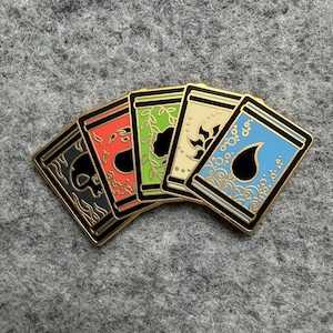 May include: Five enamel pins with gold borders, each featuring a different design: a skull, a flame, leaves, a sun, and water droplets. The pins are in a row, with a gray background.