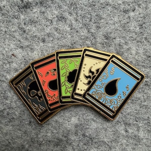 Könnte beinhalten: Eine Sammlung von fünf Emaille-Anstecknadeln mit goldenen Rändern, jede mit einem einzigartigen Design. Die Anstecknadeln zeigen einen Totenkopf, ein Herz, Blätter, florale Muster und einen Tropfen, alle vor unterschiedlich farbigen Hintergründen.