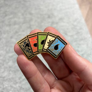 May include: A set of five enamel pins featuring card designs with gold borders. Each pin has a different color scheme: black, red, green, cream, and blue. The designs include skulls, leaves, and ornate patterns.