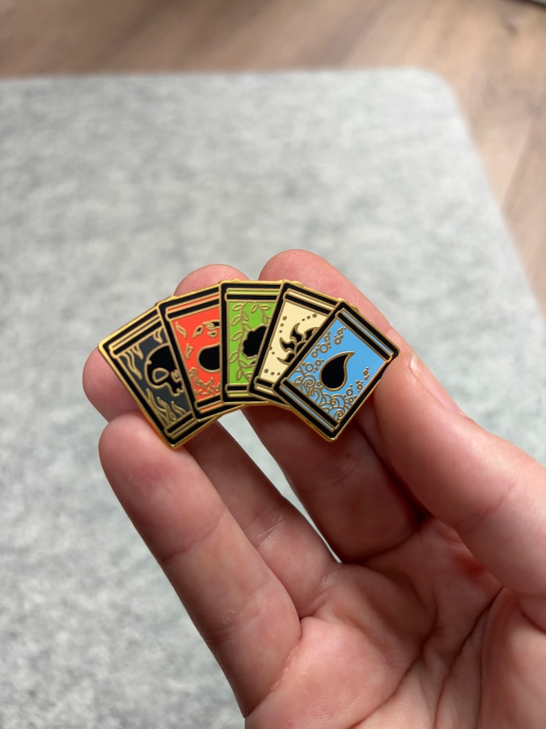 May include: A set of five enamel pins featuring card designs with gold borders. Each pin has a different color scheme: black, red, green, cream, and blue. The designs include skulls, leaves, and ornate patterns.