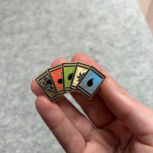 May include: A set of five enamel pins featuring card designs with gold borders. The pins display various symbols in black, red, green, yellow, and blue. The pins are held in a hand against a neutral background.