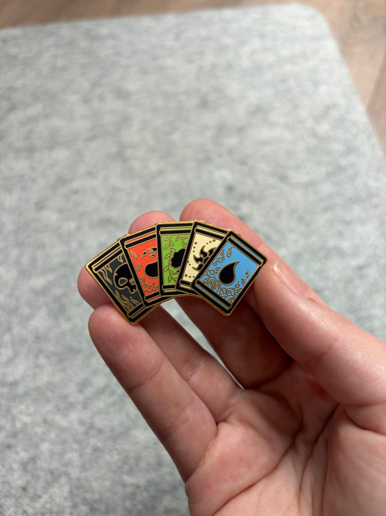 May include: A set of five enamel pins featuring card designs with gold borders. The pins display various symbols in black, red, green, yellow, and blue. The pins are held in a hand against a neutral background.
