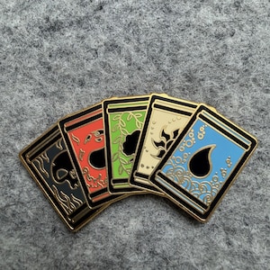 May include: Five enamel pins with gold trim, each featuring a different design. The pins are arranged in a fan shape, showcasing a skull, a stylized flame, a leafy design, a floral pattern, and water droplets. The pins have black, red, green, cream, and blue backgrounds.