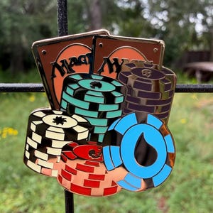 May include: A detailed pin featuring stacks of poker chips in various colors, including black, red, blue, and teal, with two cards in the background. The cards have the word "Magic" on them. The pin has a gold-colored border.