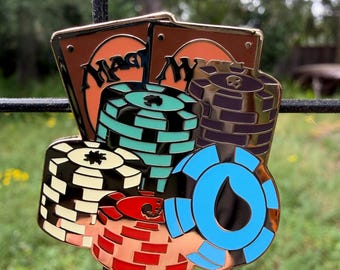 May include: A detailed pin featuring stacks of poker chips in various colors, including black, red, blue, and teal, with two cards in the background. The cards have the word "Magic" on them. The pin has a gold-colored border.
