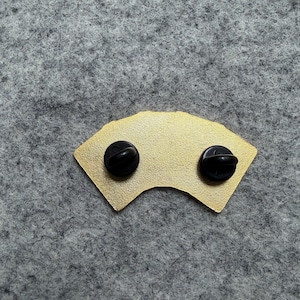 May include: A gold-colored pin in the shape of a fan, with two black pin backs. The pin has a smooth, metallic finish and is set against a gray background.