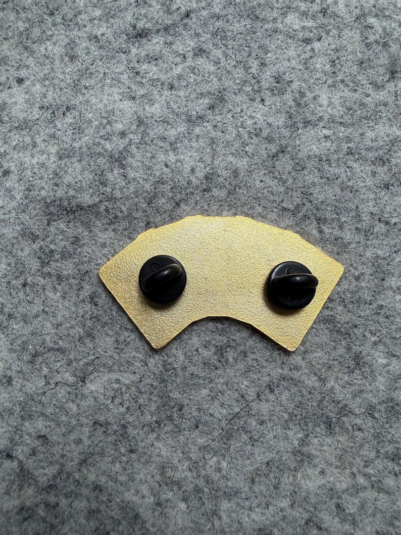 May include: A gold-colored pin in the shape of a fan, with two black pin backs. The pin has a smooth, metallic finish and is set against a gray background.
