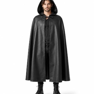 May include: A floor-length black hooded cape featuring a smooth, leather-like finish. The garment includes a deep hood, a structured shoulder yoke, and a simple tie closure at the neck. The cape hangs in a wide, open silhouette over a black shirt.